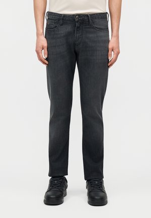 Άτομο που φοράει τζιν σκουρόχρωμο γκρι με ευθεία γραμμή και μαύρα αθλητικά παπούτσια, στέκεται με τα χέρια χαλαρά στο πλάι, μπροστά σε απλό φόντο.