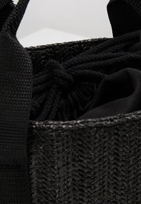 Bolsa de tote tejida negra con un exterior texturizado, forro de tela negra y cierre de cordón. Asas hechas de material resistente.