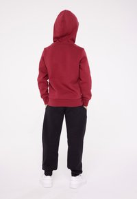 Felpa rossa con cappuccio regolabile, polsini aderenti e tasca a marsupio, abbinata a pantaloni sportivi neri. Scarpe da ginnastica bianche completano l'outfit.