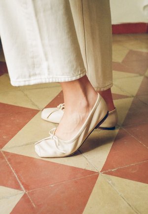 Pėdos dėvinčios kreminės spalvos kvadratinės priekinės dalies baleto batelius su mažais kaspinėliais, stovinčios ant smėlio ir terakotos rašto plytelių grindų.