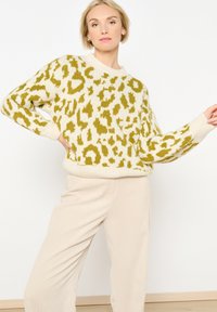 Sweter w desenie lamparta w kolorze kremowym i zielonym, z okrągłym dekoltem, ściągaczami przy rękawach oraz luźnym krojem. Zestawiony z beżowymi spodniami z kordurowej tkaniny.