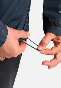 Marine jasje met een gestructureerde oppervlakte, met een toggelaar die door een hand wordt aangetrokken; klittenband manchet detail zichtbaar.