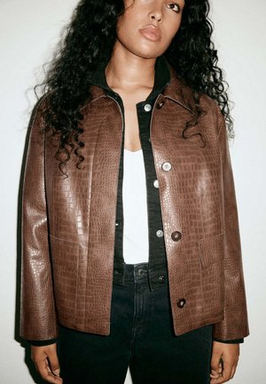Frau mit lockigem Haar trägt eine braune Lederjacke mit Krokodilmuster über einem schwarzen Hemd mit Knöpfen und einem weißen Top, stehend vor neutralem Hintergrund.