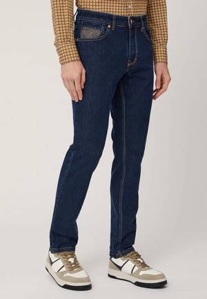 Jeans in denim indaco con vestibilità slim, cuciture arancioni a contrasto e cinque tasche. Abbinati a sneaker bianche e beige.