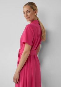 Fuchsia mouwjurk met een kraag en tailleband, met een vloeiende stijl en geplooide textuur. Het model staat met de rug naar de camera, waardoor de achterkant mooi zichtbaar is.