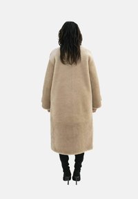 Beige shearlingjacka med lös passform, midja-l�ängd, vida ärmar och en slät yta, sedd från baksidan. Svarta högklackade stövlar synliga.