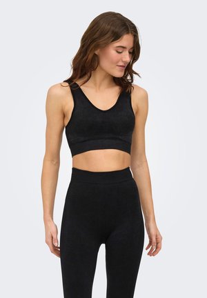 Kobieta ubrana w czarny bezrękawnik typu crop top i legginsy z wysokim stanem, stojąca z zamkniętymi oczami i lekko opuszczoną głową.