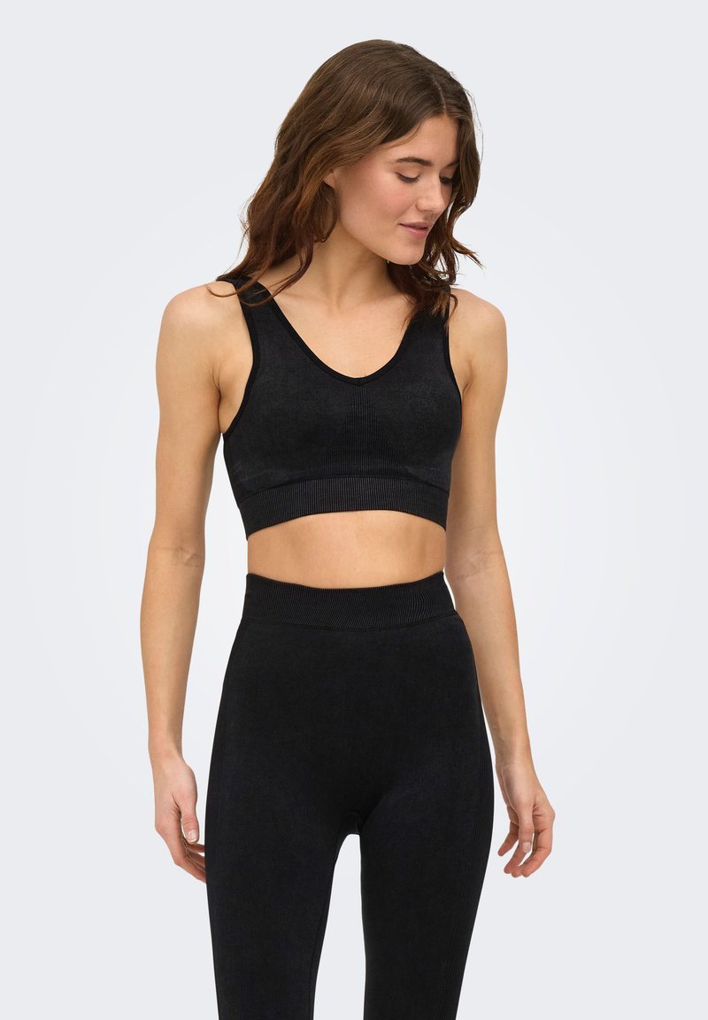 Γυναίκα που φοράει μαύρο αμάνικο crop top και κολάν με ψηλή μέση, στέκεται με κλειστά μάτια και ελαφρώς σκυμμένο κεφάλι.