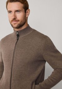 Muž s krátkou bradou v hnedé mikine so zipsom, s rukou v vrecku, čelí doľava na jednoliatom pozadí.