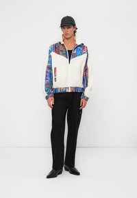Multicolored zip-up jacket with a fluffy white body, patterned sleeves, elastic cuffs, and pockets, paired with black tapered pants and shoes.