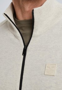 Beige gestrickter Reißverschluss-Pullover mit einem schwarzen Reißverschluss, geripptem Kragen und einem cremefarbenen Etikett mit erhabener Schrift und Logo.