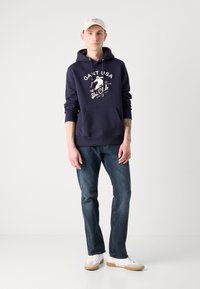 Sudadera con capucha navy con estampado gráfico blanco, bolsillo frontal, combinada con jeans azules y zapatillas blancas, con una gorra beige, diseño minimalista.