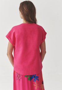 Top a maniche corte di un brillante colore rosa, realizzato in un tessuto morbido, con orlo arrotondato e vestibilità rilassata, indossato sopra una gonna dai colori vivaci con fantasia floreale.