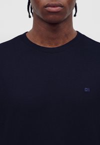 Close-up af en person med flettet hår iført en ensfarvet mørk marineblå rundhalset sweater med et lille broderet logo på brystet.