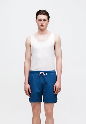 Jonge man draagt een wit tanktop en blauwe zwembroek met een wit trekkoord, staand tegen een effen witte achtergrond.