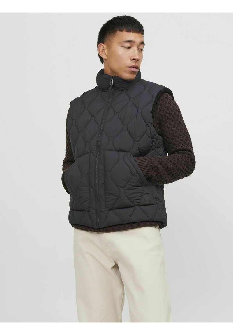 Schwarze gesteppte Weste mit hohem Kragen, Reißverschluss und zwei Seitentaschen, über einem strukturierten dunkelbraunen Pullover getragen. Helle Hose.