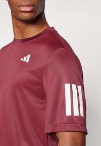 Camiseta deportiva color marrón de tela lisa, con un logo blanco en el pecho y tres rayas blancas en la manga izquierda.