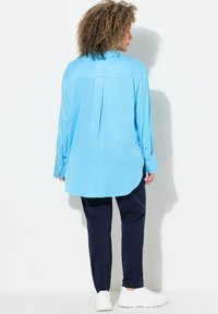 Camicia a maniche lunghe azzurro chiaro con piega sul retro, orlo curvo e polsini con bottone, abbinata a pantaloni blu navy e sneakers bianche.