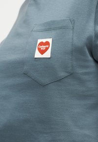 Primo piano di una tasca di camicia blu con una toppa bianca che presenta un cuore rosso e la scritta "Carhartt WIP".