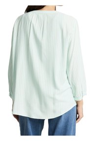Femme portant une blouse à rayures vert clair à manches longues, associée à un jean bleu, vue de dos sur un fond blanc.