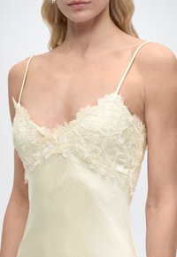 Γυναίκα που φοράει κρεμ σατέν φόρεμα slip με λεπτό δαντελένιο τελείωμα και λεπτούς ιμάντες στους ώμους, εμφανίζεται ο πάνω κορμός και ο λαιμός.