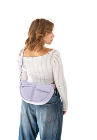 Ljuslila axelväska med halvmåneform, vadderad textur och framficka. Bärs som crossbody över en vit ribbad tröja och jeans.