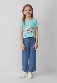 Lichtblauwe t-shirt met een kleurrijke sterrenafbeelding, gecombineerd met losse denim broek en witte sneakers. Het ontwerp bevat tekst rond de ster.