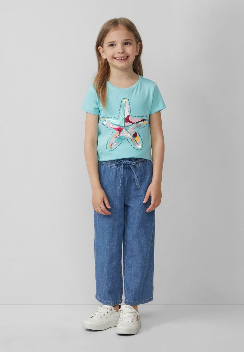 Lichtblauwe t-shirt met een kleurrijke sterrenafbeelding, gecombineerd met losse denim broek en witte sneakers. Het ontwerp bevat tekst rond de ster.