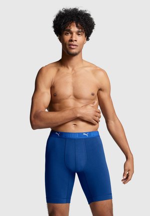 Oberkörperfreier Mann mit lockigem Haar, der blaue Puma-Boxershorts trägt und vor einem schlichten Hintergrund mit einer Hand über der Brust steht.