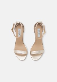 Kultaiset korkokenkä sandaalit laajalla etuhihnalla ja säädettävällä nilkkahihnalla, joissa on sileä metallipinta ja pehmustettu pohjallinen.