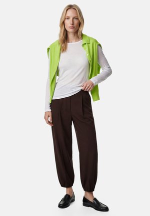 Mujer con camiseta blanca de manga larga, chaleco sin mangas verde brillante con botones, pantalones sueltos marrón oscuro y mocasines negros, de pie frente a un fondo liso.