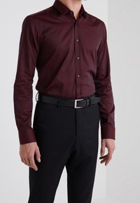 Camisa burdeos de manga larga con cuello abotonado, que presenta una textura suave y botones negros en contraste, combinada con pantalones y cinturón negros.