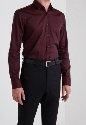 Camisa burdeos de manga larga con cuello abotonado, que presenta una textura suave y botones negros en contraste, combinada con pantalones y cinturón negros.