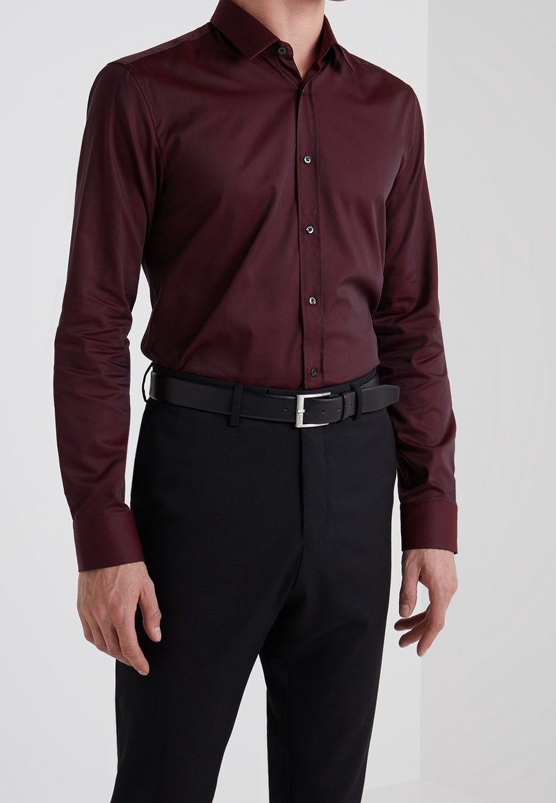 Camisa burdeos de manga larga con cuello abotonado, que presenta una textura suave y botones negros en contraste, combinada con pantalones y cinturón negros.
