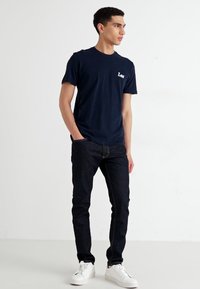 Marine bomull t-skjorte med hvit "Lee" logo, kombinert med mørk denim jeans og hvite sneakers, presentert mot en enkel bakgrunn.
