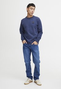 Navy crewneck sweatshirt with a patch, paired with blue straight-leg jeans and cream sneakers with brown accents. Smooth fabric texture.