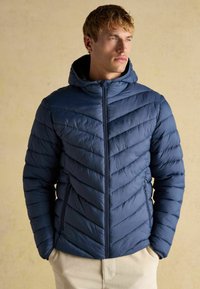 Marineblå pufferjakke med et chevronmønster, polstret design og en aftagelig hætte. Har lynlåslommer i siden og en lynlåslukning foran.