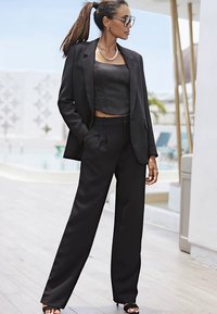 Svart skräddarsydd kostym med vida byxor och en kort topp. Har en strukturerad blazer och ett kraftigt kedjehalsband. Slät tygstruktur.
