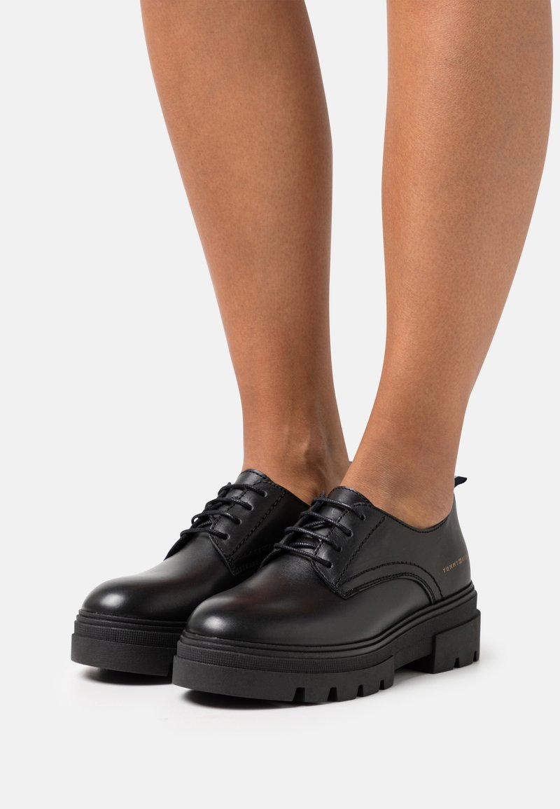 Chaussures à lacets en cuir noir avec un bout arrondi, une semelle épaisse noire et volumineuse, et des coutures décoratives minimalistes. Présentent des accents de marque.