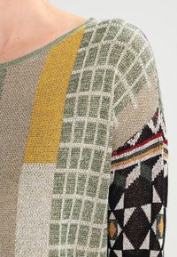 Pullover lavorato a maglia con motivi geometrici verdi, gialli e beige, filati testurizzati e una manica nera con accenti di diamanti colorati.