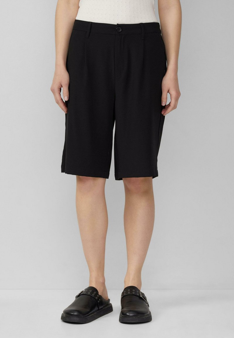 Schwarze maßgeschneiderte Shorts mit flacher Vorderseite, Knopfverschluss und seitlichen Taschen, die bis zur Mitte des Oberschenkels reichen. Glatter Stoff mit sauberen Abschlüssen.