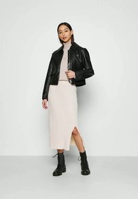 Jaqueta cropped de couro preta com gola, combinada com um vestido midi bege com texturização e fenda lateral, acompanhado de botas pretas com atacadores.