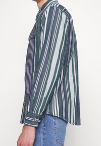 Camisa de rayas en verde y azul marino, con líneas verticales. Está hecha de una tela ligera y cuenta con un bolsillo en el pecho con botones y mangas con puños.