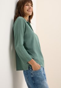 Maglia verde a maniche lunghe con collo rotondo e maniche con polsini, abbinata a jeans in denim blu. Il tessuto sembra morbido con una vestibilità rilassata.