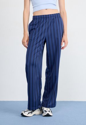 Persoon draagt losse blauwe broek met verticale strepen, witte sneakers en een lichtblauw crop top, staand tegen een effen achtergrond.