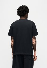 Μαύρο βαμβακερό T-shirt με κοντά μανίκια και κλασικό στρογγυλό λαιμόκοψη. Λείο ύφασμα και χαλαρή εφαρμογή, εμφα νιζόμενο από πίσω.