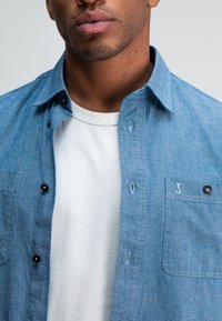 Lichtblauw chambray overhemd met zwarte knopen, een linker borstzak en borduring. Een wit T-shirt met ronde hals is zichtbaar eronder.