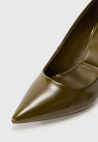 Talons hauts en cuir verni vert olive, bout pointu, surface lisse, design décolleté avec une ouverture arrondie sur le côté.