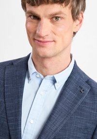 Dunkelblau gemusterte Blazer mit einem hellblauen Hemd darunter. Auffälliges silbernes Hardware-Detail am Revers des Blazers.