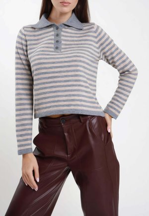 Donna che indossa una polo a maniche lunghe a righe grigie e beige e pantaloni in pelle marrone scuro a vita alta, con una mano sul fianco.
